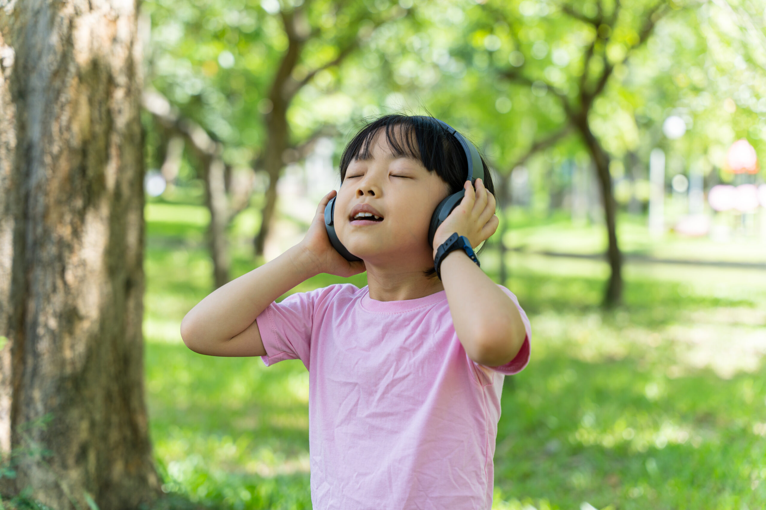 Portrait of child girl listens to music with modern headphones in park outdoors. Happy child enjoying rhythms in listening to music with headphones wireless